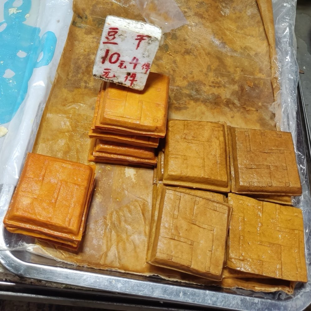 A market tray of golden-brown dougan squares beside a handwritten 豆干 price sign