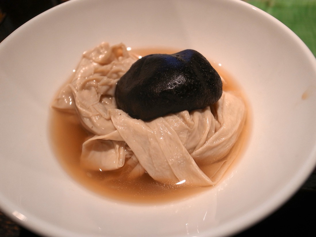 A bowl of soft folded yuba in broth, topped with a black shiitake mushroom